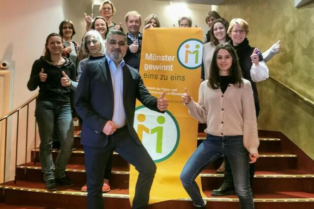 Gruppenfoto von Menschen, die vor einem Banner mit der Aufschrift „Münster gewinnt eins zu eins“ stehen und Daumen hoch zeigen.