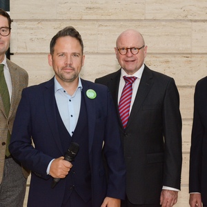 Vier Männer in formeller Kleidung posieren gemeinsam vor einer hellen Steinwand, einer hält ein Mikrofon in der Hand.
