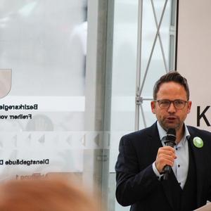 Mann mit Brille spricht am Mikrofon, im Hintergrund ein Transparent mit dem Wort „Kompetenz“.