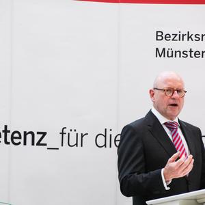 Mann in Anzug spricht an einem Rednerpult vor einem Banner der Bezirksregierung Münster.