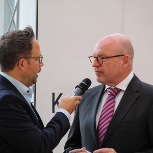 Zwei Männer führen ein Interview; einer hält ein Mikrofon, der andere hört zu, vor einer hellen Wand.