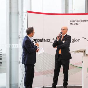 Zwei Personen stehen vor einem Rednerpult, während eine Präsentation zur Bezirksregierung Münster stattfindet.