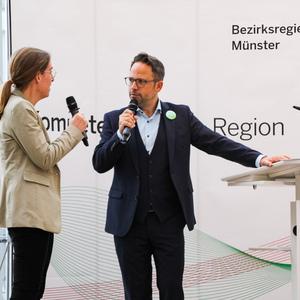 Zwei Personen sprechen auf einer Veranstaltung der Bezirksregierung Münster, Mikrofone in der Hand, Hintergrund mit Plakat.