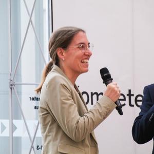 Zwei Personen sprechen miteinander, eine Frau mit Mikrofon in beige Jacke, der Mann trägt Anzug und lächelt.