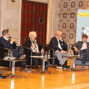 Diskussionsrunde mit fünf Personen auf einem Podium, umgeben von dekorativen Wänden und einem Banner im Hintergrund.