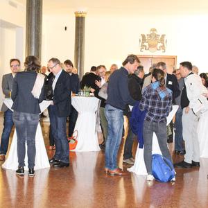 Gruppengespräch auf einer Veranstaltung in einem großen, hellen Saal mit Stehtischen und Teilnehmern in formeller Kleidung.