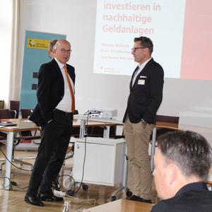 Zwei Herren im Anzug stehen in einem Konferenzraum, im Hintergrund eine Präsentation zu nachhaltigen Geldanlagen.
