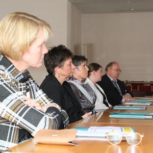 Teilnehmer einer Besprechung sitzen an einem Tisch. Dokumente und Wasserflaschen sind sichtbar.