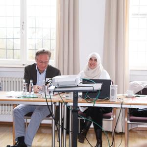 Drei Personen sitzen an einem Tisch, an dem ein Laptop und ein Projektor stehen, während sie in einem hellen Raum arbeiten.