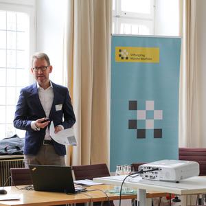 Mann in einem Anzug hält ein Präsentationsgerät, steht vor einem Banner und einem Laptop in einem Konferenzraum.