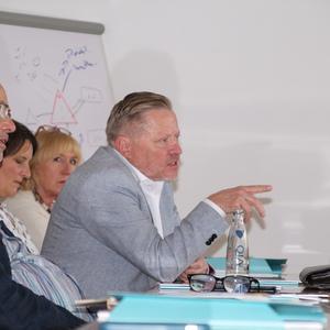 Eine Gruppe von Personen in einem Meeting, der Mann in Grau spricht und gestikuliert, während andere zuhören.