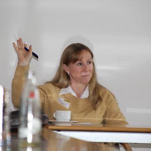 Frau mit blonden Haaren hebt die Hand und hält einen Stift, während sie an einem Tisch in einem Konferenzraum sitzt.