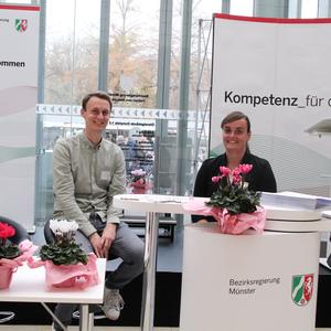 Zwei Personen lächeln am Info-Stand der Bezirksregierung Münster, umgeben von Blumen und Informationsmaterial.