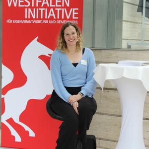 Frau mit Locken und Brille sitzt auf einem Hocker vor einem roten Banner der Stiftung Westfalen Initiative.