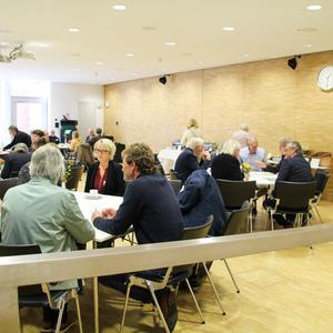 Gruppengespräch mehrerer Personen an Tischen in einem hellen, modernen Raum mit Pflanzen und einer Uhr an der Wand.