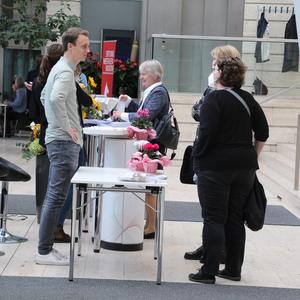 Menschen stehen um Tische in einer modernen Umgebung, diskutieren und tauschen Informationen aus. Blumen in Töpfen dekorieren die Tische.