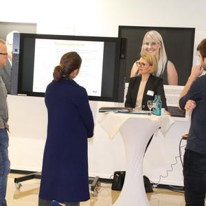 Vier Personen diskutieren an einem Stehtisch vor einem Monitor mit Präsentation und Grafiken.