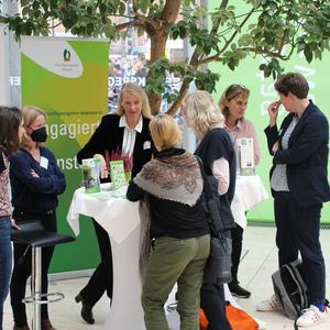 Menschen stehen an einem Stehtisch, umgeben von Informationsmaterialien, in einem hellen Raum mit Pflanzen.