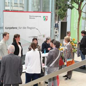 Menschen stehen an einem Informationsstand der Bezirksregierung Münster und diskutieren. Im Hintergrund Pflanzen und ein Rednerpult.