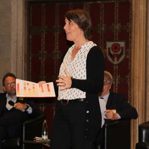 Frau spricht auf einer Bühne und hält ein Papier, während zwei Männer im Hintergrund sitzen.