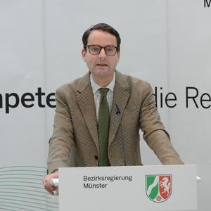 Mann im karierten Anzug steht an einem Rednerpult der Bezirksregierung Münster mit Hintergrundbeschriftung „Kompetent für die Region“.
