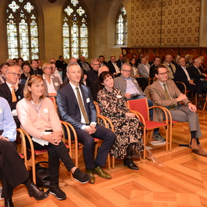 Gruppe von Menschen sitzt im Präsentationssaal, angestrengt auf die Bühne blickend, mit historischen Fenstern im Hintergrund.