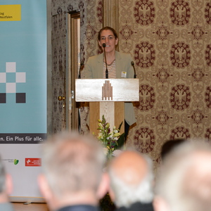 Frau am Rednerpult, vor einem Banner mit dem Schriftzug „Stiftungstag Münster Westfalen“, im Hintergrund Teilnehmer.