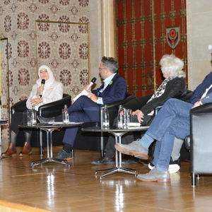 Podiumsdiskussion mit sechs Personen, Mikrofone und Wassergläser auf Tisch, hintergrund mit dekorativen Mustern.