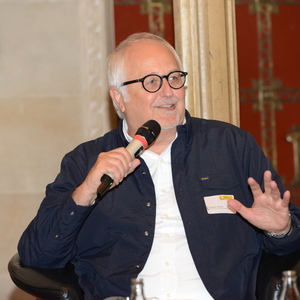 Mann mit Brille hält ein Mikrofon und spricht in einem Forum, braune Wände im Hintergrund.