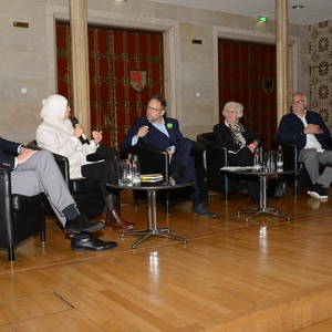 Podiumsdiskussion mit mehreren Rednern in schwarzen Sesseln, auf einer Bühne mit Holzfußboden und historischen Wandverzierungen.