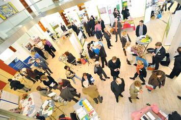 Überblick über eine Messe mit vielen Besuchern, Ständen und Informationsmaterialien in einem hellen, modernen Raum.