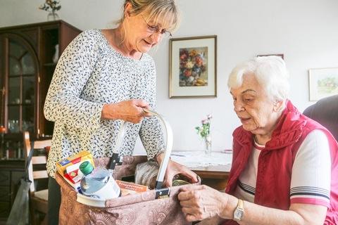 Zwei Frauen in einem Wohnraum schauen in einen Einkaufskorb, gefüllt mit Lebensmitteln und Getränken.