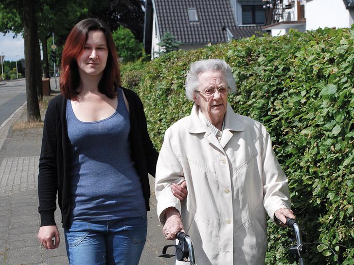 Eine junge Frau unterstützt eine ältere Dame mit Rollator beim Spazieren auf einem Gehweg, neben einer Hecke.