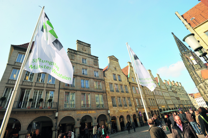 Flaggen mit der Aufschrift „Stiftungstag Münster-Westfalen“ wehen vor historischen Gebäuden und Passanten auf einem Platz.