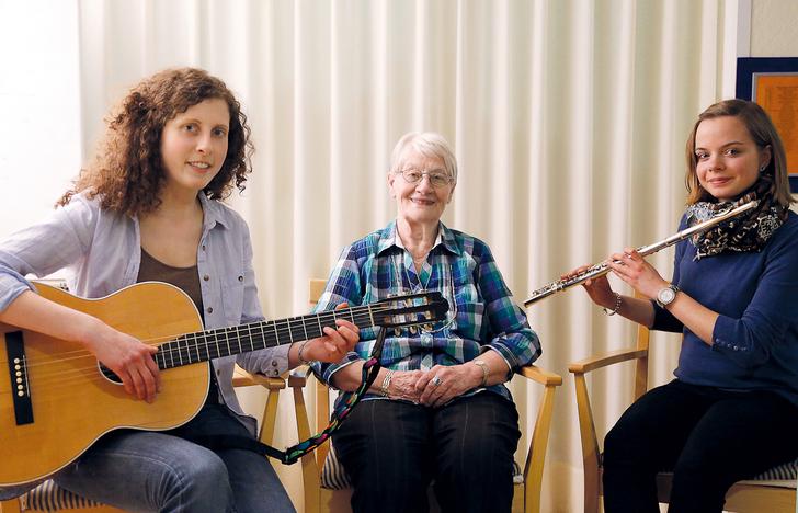 Drei Frauen sitzen in einem Raum: eine mit Gitarre, eine mit Fl&ouml;te und eine l&auml;chelnde &auml;ltere Frau dazwischen.