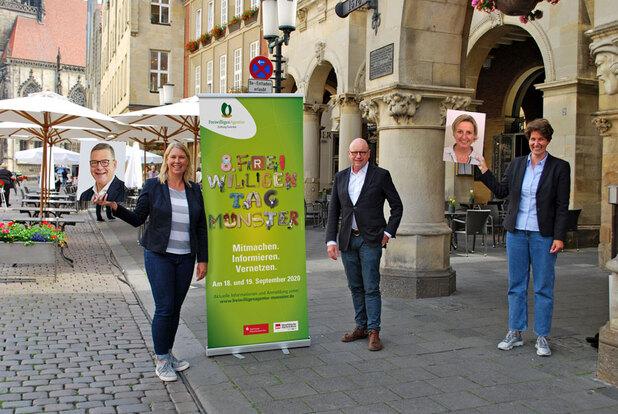 Gruppenszene auf einer Straße mit einer Informationswand über den Freiwilligentag in Münster und Personen mit Bildporträts.