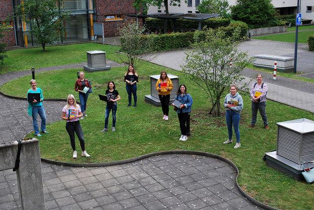 Gruppenfoto von zehn Frauen im Freien, einige halten Bücher, umgeben von Bäumen und modernen Gebäuden.