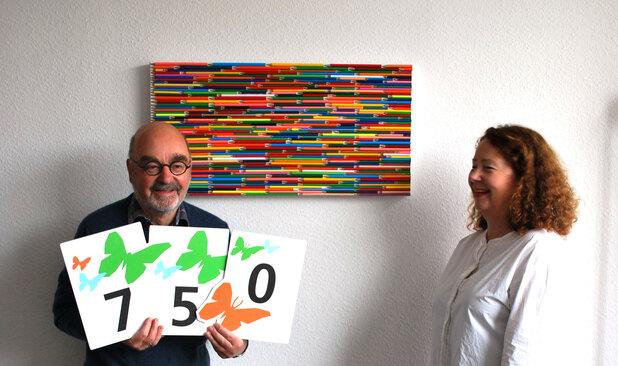 Ein Mann hält drei Karten mit Zahlen und Schmetterlingen, neben ihm steht eine lächelnde Frau vor einer bunten Wanddekoration.