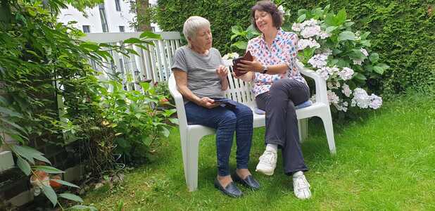 Zwei Frauen sitzen auf einer Bank im Garten und betrachten gemeinsam ein Tablet, umgeben von Pflanzen und Blumen.