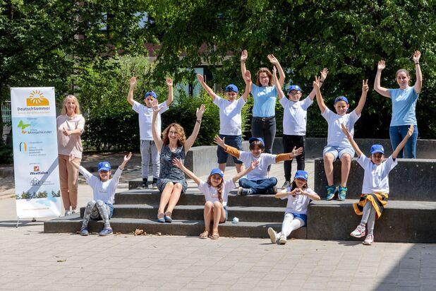 Gruppe von Kindern und Betreuern posiert fröhlich vor einem Banner des Projekts „DeutschSommer“ auf Stufen im Freien.