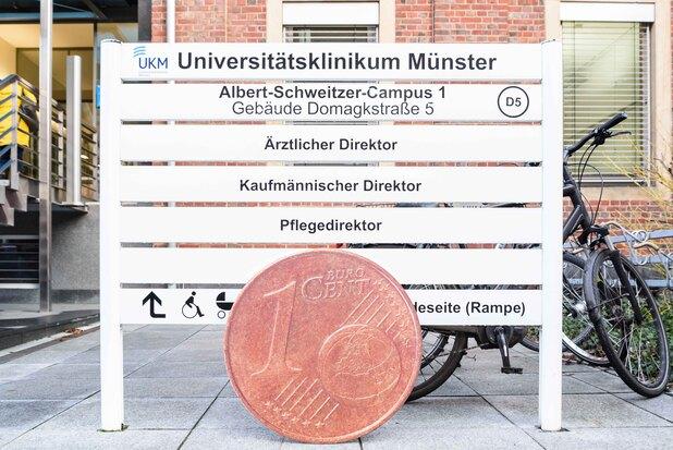 Nahaufnahme eines 1-Cent-Münze vor einem Richtungsschild des Universitätsklinikums Münster mit Fahrrädern im Hintergrund.