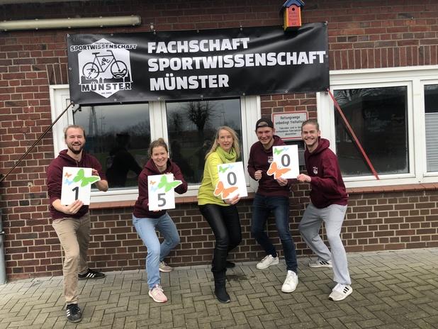 Fünf Personen in Sweatshirts halten Zahlenkarten mit 1, 5 und 0 vor einem Banner der Fachschaft Sportwissenschaft Münster.