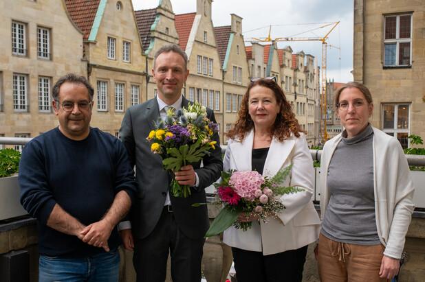 Vier Personen stehen in einem Innenhof, zwei halten Blumensträuße, im Hintergrund sind historische Gebäude sichtbar.