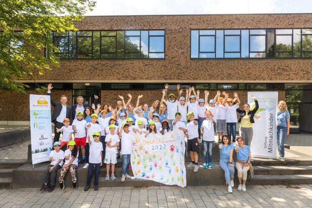Gruppenszene von Kindern und Erwachsenen vor einem Gebäude, alle lächeln und winken, mit einem bunten Banner in der Mitte.