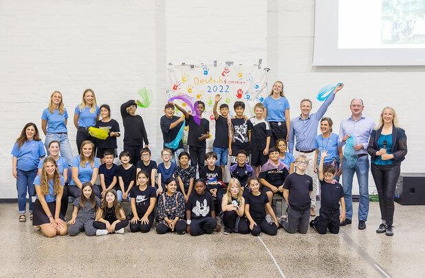 Gruppe von Kindern und Erwachsenen bei einer Feier mit bunten Tüchern, vor einem Wandplakat mit Handabdrücken.