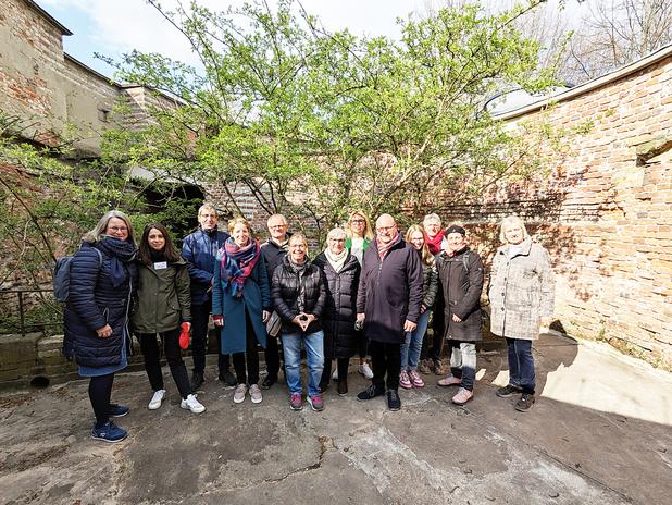 Gruppe von zwölf Personen posiert vor einer Backsteinmauer mit einem Baum in einem begrünten Innenhof.