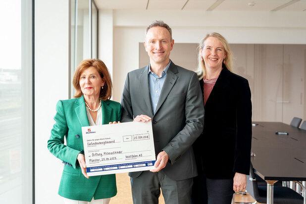 Drei Personen präsentieren einen Scheck über 25.000 Euro für die Stiftung Mitmachkinder in einem modernen Raum.