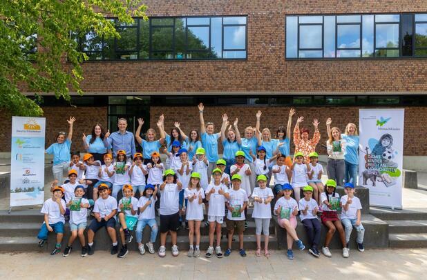 Gruppenfoto von Kindern und Betreuern mit erhobenen Händen vor einer Schule, umgeben von Werbebannern für „DeutschSommer“.