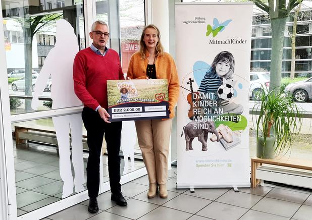 Zwei Personen halten einen Scheck über 2000 Euro vor einem Banner der Stiftung MitmachKinder in einem hellen Raum.