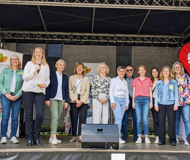Gruppe von Frauen und Männern lächelt auf einer Bühne, Mikrofon und Präsentationsmaterial im Hintergrund sichtbar.